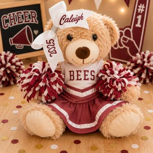 May include: A tan plush teddy bear wearing a white and maroon cheerleader uniform with the word BEES printed on the chest. The bear has a white hair bow featuring the name Caleigh and the year 2025, and holds two maroon and white pom-poms.