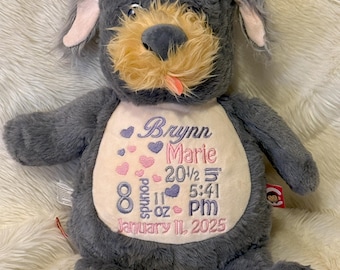 May include: A plush gray dog toy with a tan muzzle and a pink tongue. The chest features embroidered text: Brynn Marie, 20 1/2 inches, 8 pounds 11 ounces, 5:41 PM, and January 11, 2025, accented with small purple and pink heart-shaped embroidery.