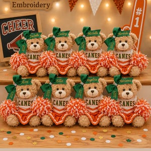 May include: Eight light brown plush teddy bears dressed as cheerleaders in white and orange uniforms with the word CANES printed on the front. Each bear wears a green hair bow with the year 2025 and a personalized name, holding orange and white pom-poms.