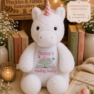 May include: A white plush unicorn toy with a pink horn, featuring embroidered text that reads Jessica's reading buddy above an illustration of an open book with flowers and butterflies. The toy has black eyes and pink nostrils.