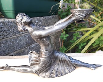 May include: A silver-toned art deco figurine of a woman in a flowing skirt, holding a small bowl aloft. The figurine is in a seated position with one leg extended.