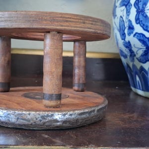 Könnte beinhalten: Ein kleiner, runder Pflanzenständer aus Holz mit vier kurzen Beinen. Der Ständer hat eine dunkelbraune Oberfläche mit einem silbernen Metallband um die Basis. Im Hintergrund befindet sich eine blau-weiße Zier-Vase.