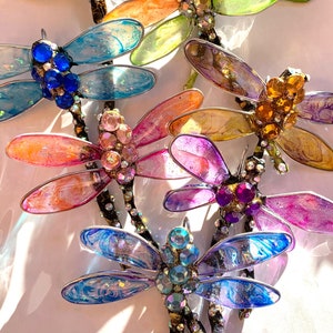 May include: A collection of eight colorful dragonfly ornaments with iridescent wings and jeweled accents. The dragonflies are arranged in a cluster, with their wings overlapping.