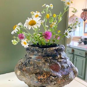 May include: A textured, rounded vase crafted with embedded stones and a dark, metallic-flecked finish. The vase contains a bouquet of wildflowers, including white daisies with yellow centers and small, vibrant pink clover blossoms on green stems.