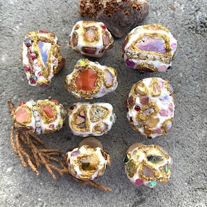 May include: Nine white rings with gold accents and various colorful stones and crystals. The rings are arranged in a circle on a gray textured surface.