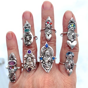 K&ouml;nnte beinhalten: Ein Handmodell tr&auml;gt acht silberne Ringe mit religi&ouml;sen Figuren und bunten Edelsteinen. Die Ringe sind alle in verschiedenen Stilen gehalten und zeigen verschiedene Heilige und religi&ouml;se Symbole.