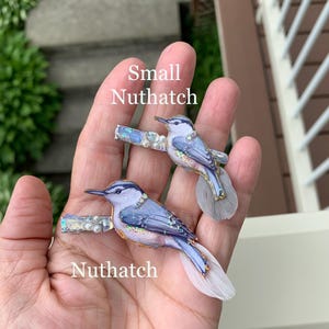 May include: Two hair clips featuring illustrations of Nuthatch birds. The birds have blue and grey plumage with iridescent glitter accents and small white pearls. Each clip is finished with a soft, light-colored feather tail. Text reads Small Nuthatch and Nuthatch.