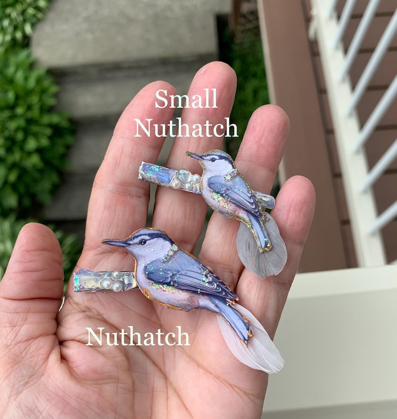 May include: Two hair clips featuring illustrations of Nuthatch birds. The birds have blue and grey plumage with iridescent glitter accents and small white pearls. Each clip is finished with a soft, light-colored feather tail. Text reads Small Nuthatch and Nuthatch.