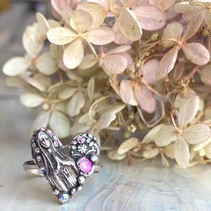 May include: A silver heart ring with intricate detailing and iridescent gemstones. The ring has a smaller, decorated oval shape and a pink gemstone. Dried flowers in soft pink and cream tones are in the background.
