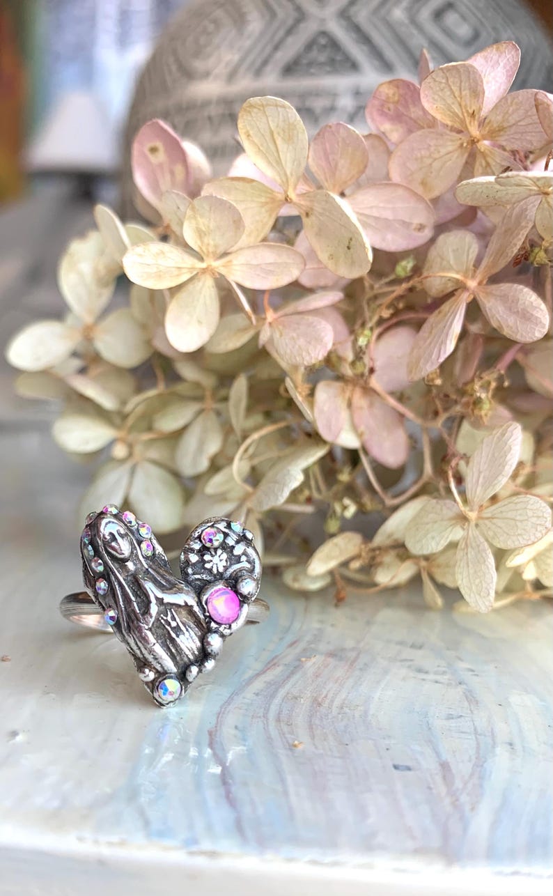 May include: A silver heart ring with intricate detailing and iridescent gemstones. The ring has a smaller, decorated oval shape and a pink gemstone. Dried flowers in soft pink and cream tones are in the background.