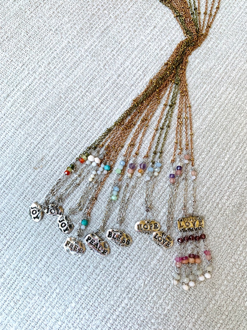 Puede incluir: Un conjunto de 12 collares de plata con diferentes cuentas de piedras preciosas y dijes. Cada dije presenta una palabra diferente, incluyendo "Alegr&iacute;a", "Paz", "Bendici&oacute;n", "Esperanza", "Amor" y "Libre".