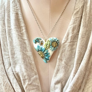Puede incluir: Un collar de cadena de plata con un colgante de coraz&oacute;n de cer&aacute;mica blanca con detalles florales azules y amarillos.
