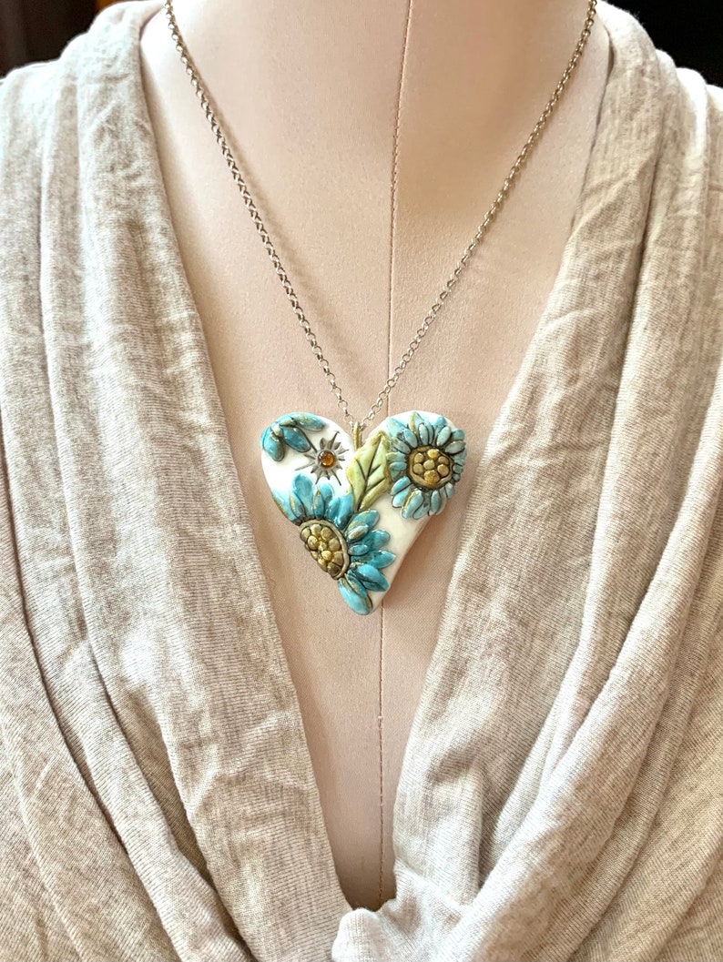 Puede incluir: Un collar de cadena de plata con un colgante de coraz&oacute;n de cer&aacute;mica blanca con detalles florales azules y amarillos.