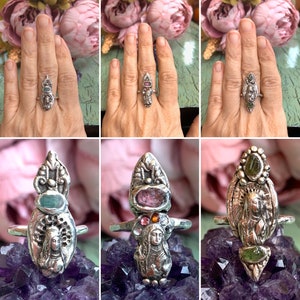 K&ouml;nnte beinhalten: Drei silberne Ringe mit einer religi&ouml;sen Figur in der Mitte, umgeben von bunten Edelsteinen. Die Ringe liegen auf einem violetten Hintergrund.