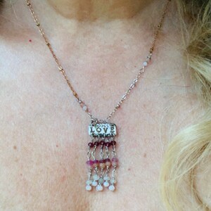 Puede incluir: Un collar de plata con un dije que dice "LOVE" y una franja de cuentas rosadas, blancas y moradas.