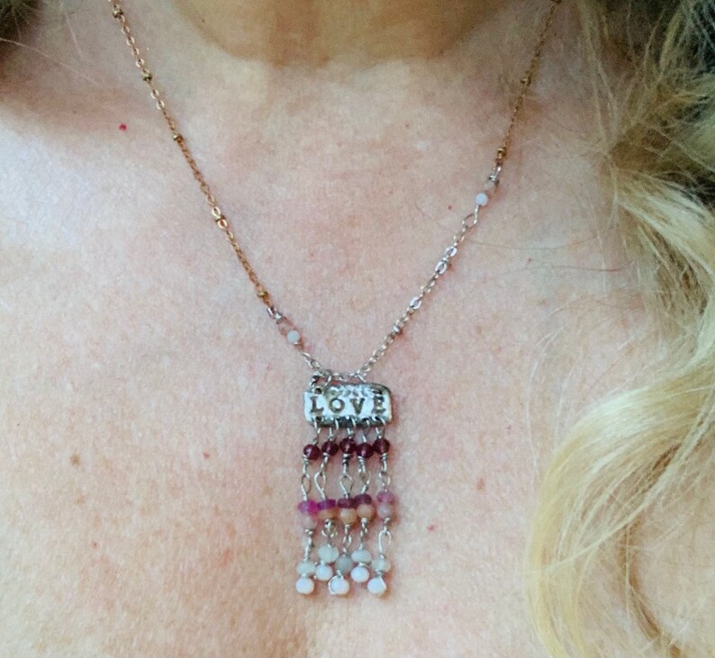 Puede incluir: Un collar de plata con un dije que dice "LOVE" y una franja de cuentas rosadas, blancas y moradas.