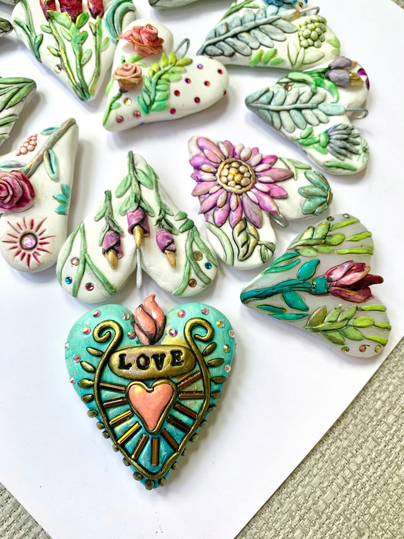 Puede incluir: Una colecci&oacute;n de adornos hechos a mano en forma de coraz&oacute;n con dise&ntilde;os florales e inspirados en la naturaleza. Un coraz&oacute;n presenta la palabra "LOVE" en letras doradas con un coraz&oacute;n rosa en su interior.