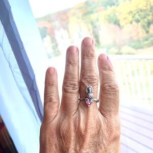 May include: A silver ring with a pink heart-shaped gemstone and a detailed figure. The ring is worn on a finger, highlighting the contrast between the silver band and the pink stone. The design is intricate and eye-catching.