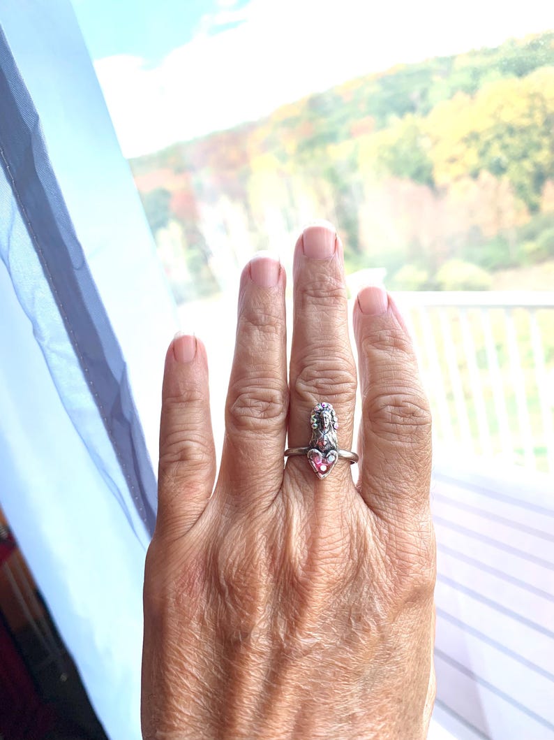 May include: A silver ring with a pink heart-shaped gemstone and a detailed figure. The ring is worn on a finger, highlighting the contrast between the silver band and the pink stone. The design is intricate and eye-catching.