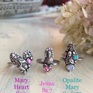 May include: Three silver rings with religious themes. One ring is heart-shaped with a Mary design, another features Jesus, and the third has an Opalite Mary design. Each ring has colourful accents and is displayed on a light-coloured surface.