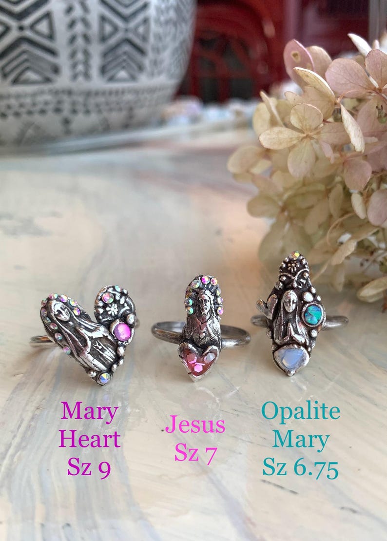 May include: Three silver rings with religious themes. One ring is heart-shaped with a Mary design, another features Jesus, and the third has an Opalite Mary design. Each ring has colourful accents and is displayed on a light-coloured surface.