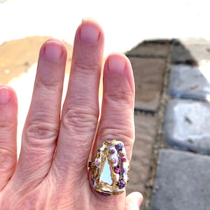 May include: A white and gold ring with a triangular shaped stone and purple accents. The ring is on a person's finger.