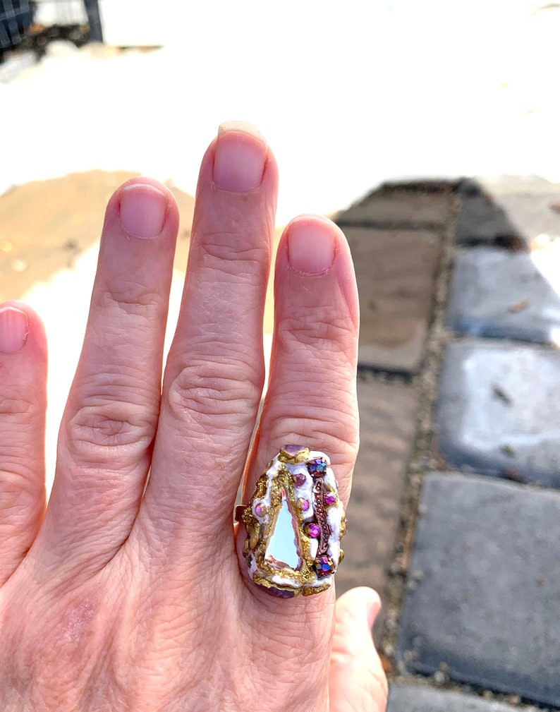 May include: A white and gold ring with a triangular shaped stone and purple accents. The ring is on a person's finger.