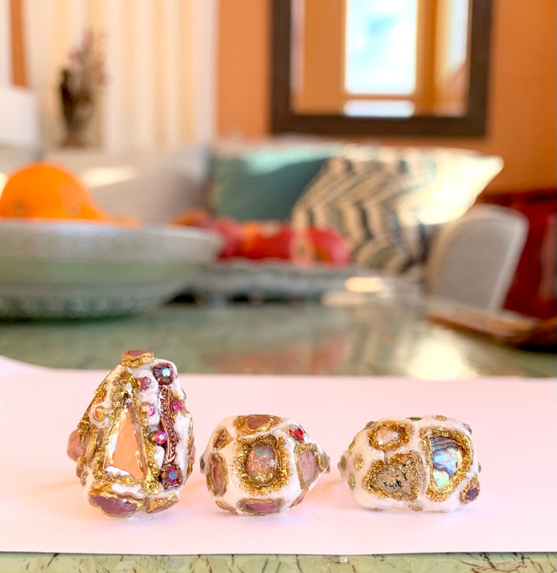 May include: Three white rings with gold and pink accents. The rings are decorated with small, colorful stones and glitter.