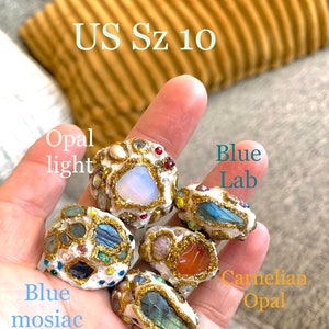 May include: Five rings made of white material with gold accents and various gemstones. The rings are labeled "Opal light", "Blue Lab", "Blue mosaic", "Carnelian Opal", and "Green Lab". The rings are on a hand, and the text "US Sz 10" is visible above the rings.