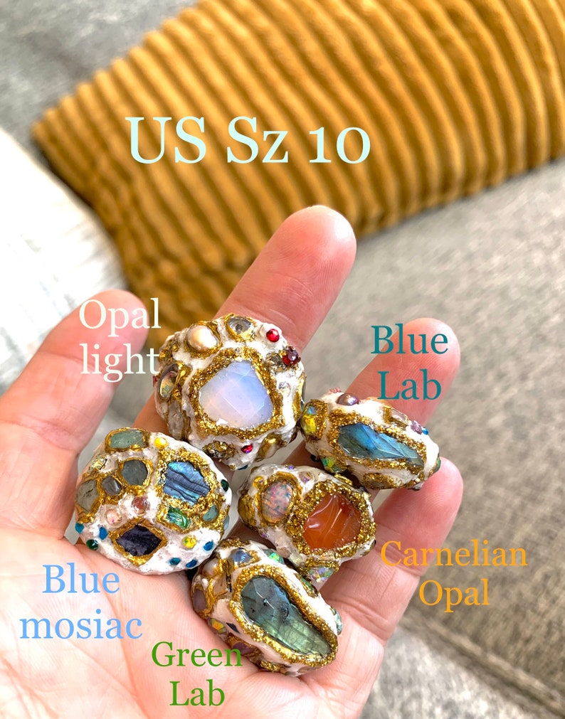 May include: Five rings made of white material with gold accents and various gemstones. The rings are labeled "Opal light", "Blue Lab", "Blue mosaic", "Carnelian Opal", and "Green Lab". The rings are on a hand, and the text "US Sz 10" is visible above the rings.