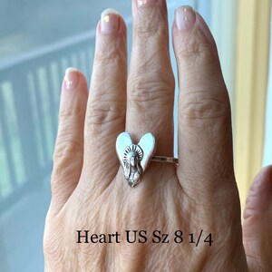 K&ouml;nnte beinhalten: Silberner Herzring mit einer religi&ouml;sen Figur im Inneren. Der Ring befindet sich an einem Finger und der Text "Heart US Sz 8 1/4" befindet sich unter dem Ring.