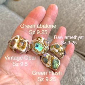 May include: Four rings with different gemstones and gold accents. The rings are made of white clay and feature green abalone, raw amethyst, vintage opal, and green flash gemstones. The rings are size 9.25 and 9.5.