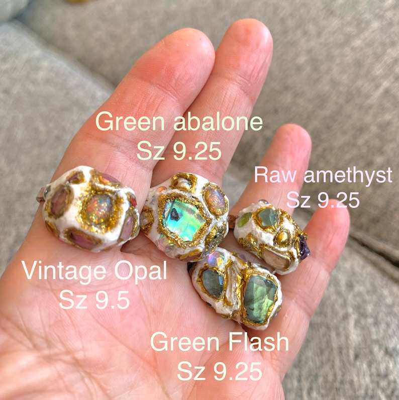 May include: Four rings with different gemstones and gold accents. The rings are made of white clay and feature green abalone, raw amethyst, vintage opal, and green flash gemstones. The rings are size 9.25 and 9.5.