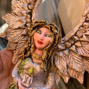 May include: A handmade angel figurine with wings made of textured white and gold leaves. The angel is holding a candle in its hands and has a light blue and green dress with gold accents.