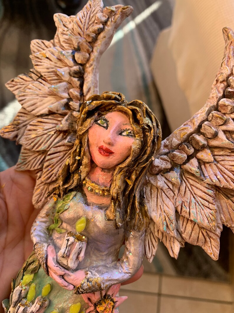 May include: A handmade angel figurine with wings made of textured white and gold leaves. The angel is holding a candle in its hands and has a light blue and green dress with gold accents.