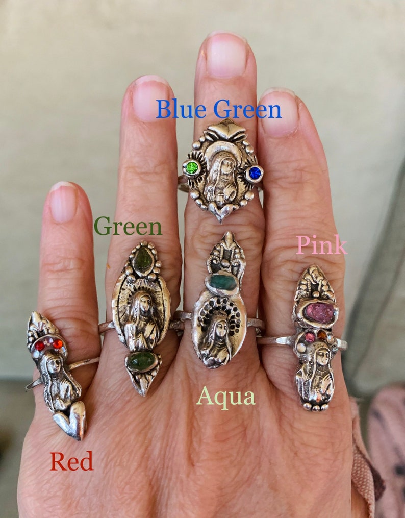 K&ouml;nnte beinhalten: F&uuml;nf Silberringe mit verschiedenen farbigen Steinen. Die Ringe zeigen eine religi&ouml;se Figur in der Mitte. Die Steine sind rot, gr&uuml;n, blau, rosa und aquamarin.