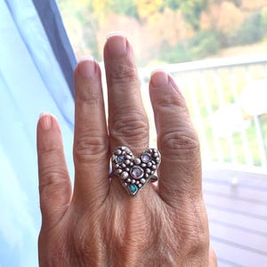 May include: A silver ring with a heart-shaped design. The ring has a textured surface with small, round embellishments and several iridescent gemstones. The ring is worn on a finger, highlighting its intricate details and craftsmanship.