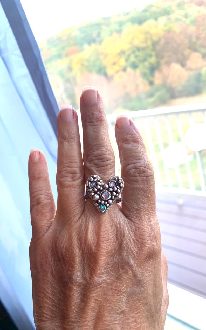 May include: A silver ring with a heart-shaped design. The ring has a textured surface with small, round embellishments and several iridescent gemstones. The ring is worn on a finger, highlighting its intricate details and craftsmanship.