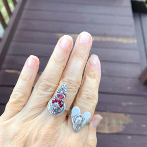 K&ouml;nnte beinhalten: Zwei silberne Ringe mit religi&ouml;sen Motiven. Ein Ring hat ein herzf&ouml;rmiges Design mit einer Darstellung der Jungfrau Maria. Der andere Ring hat einen silbernen Anh&auml;nger mit roten Edelsteinen und einer Darstellung der Jungfrau Maria.