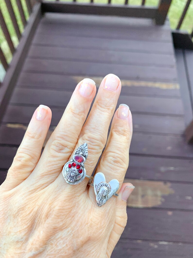 K&ouml;nnte beinhalten: Zwei silberne Ringe mit religi&ouml;sen Motiven. Ein Ring hat ein herzf&ouml;rmiges Design mit einer Darstellung der Jungfrau Maria. Der andere Ring hat einen silbernen Anh&auml;nger mit roten Edelsteinen und einer Darstellung der Jungfrau Maria.