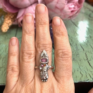 K&ouml;nnte beinhalten: Ein silberner Ring mit einem rosafarbenen und orangefarbenen Edelstein und einer detaillierten Figur einer Frau. Der Ring wird an einem Finger getragen.