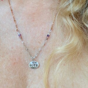 Puede incluir: Un collar de plata con un peque&ntilde;o colgante ovalado que dice "LOVE". El collar tiene una cadena delicada con peque&ntilde;as cuentas moradas.