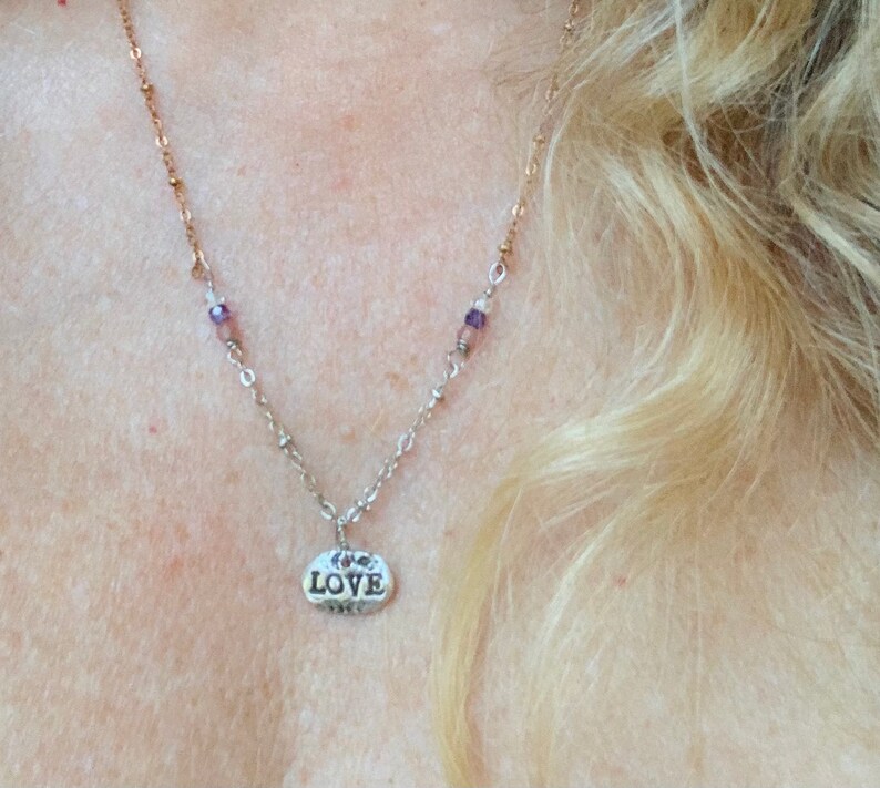 Puede incluir: Un collar de plata con un peque&ntilde;o colgante ovalado que dice "LOVE". El collar tiene una cadena delicada con peque&ntilde;as cuentas moradas.