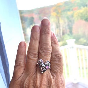 May include: A silver ring with a heart-shaped design, featuring a religious figure and pink gemstones. The ring is worn on a hand, with a scenic outdoor background visible.