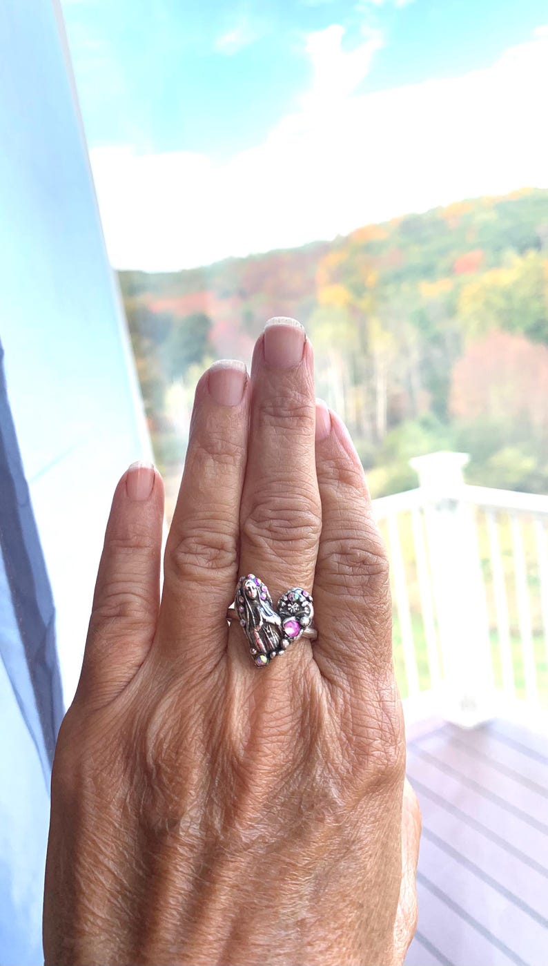 May include: A silver ring with a heart-shaped design, featuring a religious figure and pink gemstones. The ring is worn on a hand, with a scenic outdoor background visible.