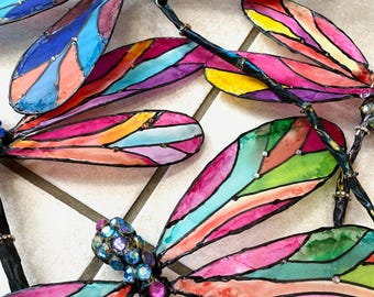 May include: Four colorful dragonfly sculptures with iridescent wings and black outlines. The wings are painted with stripes of pink, blue, green, orange, and yellow. The dragonflies are made of paper and have a wire body.