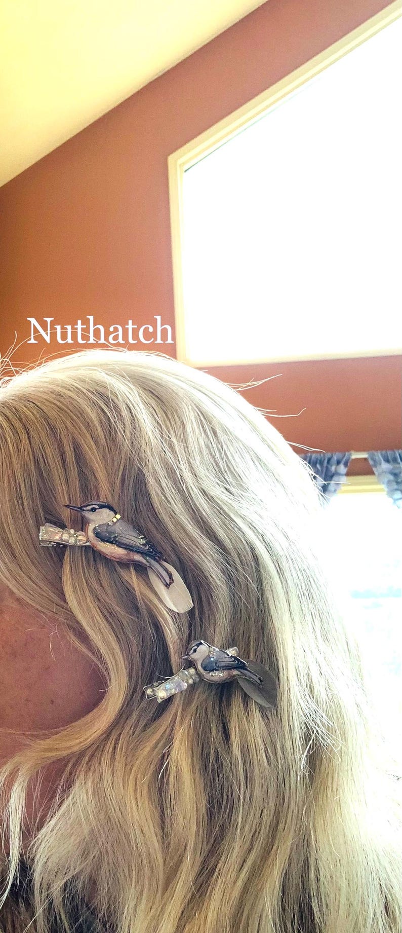 May include: Two hair clips featuring detailed Nuthatch bird figures with blue, white, and brown plumage. Each bird is attached to a metallic clip base with small, clear, reflective accents. The word Nuthatch is printed in white text on the left.
