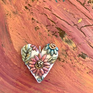 Puede incluir: Un adorno de cer&aacute;mica blanca en forma de coraz&oacute;n con un dise&ntilde;o en relieve de flores rosas, azules y verdes. El adorno est&aacute; sobre un fondo texturizado marr&oacute;n rojizo.
