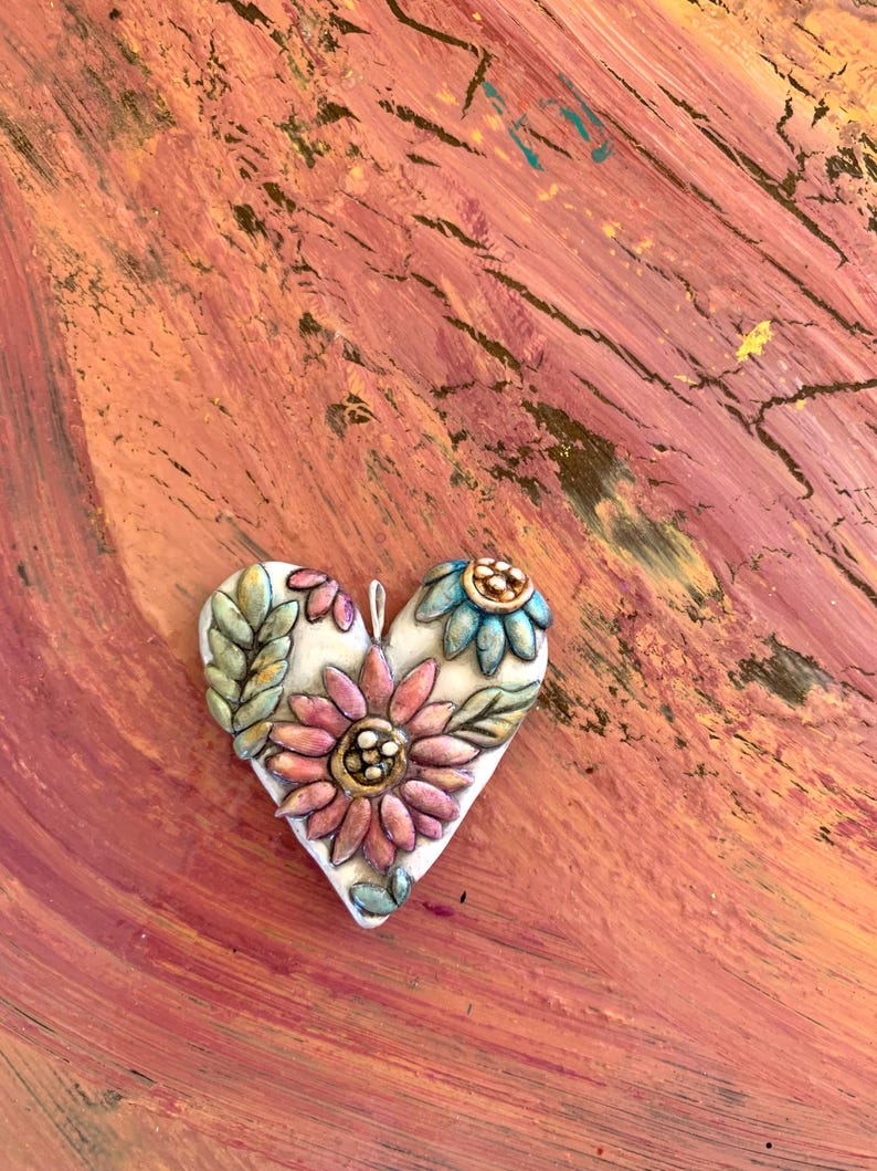 Puede incluir: Un adorno de cer&aacute;mica blanca en forma de coraz&oacute;n con un dise&ntilde;o en relieve de flores rosas, azules y verdes. El adorno est&aacute; sobre un fondo texturizado marr&oacute;n rojizo.