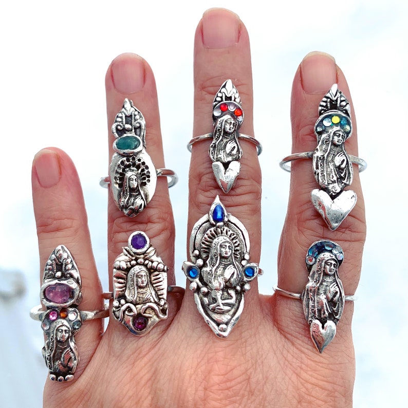 K&ouml;nnte beinhalten: Ein Handmodell tr&auml;gt acht silberne Ringe mit religi&ouml;sen Figuren und bunten Edelsteinen. Die Ringe sind alle in verschiedenen Stilen gehalten und zeigen verschiedene Heilige und religi&ouml;se Symbole.